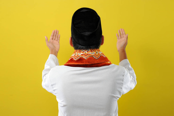 Jeune homme musulman asiatique levant la main et priant, vue de dos, isolé sur fond jaune - Photo, image