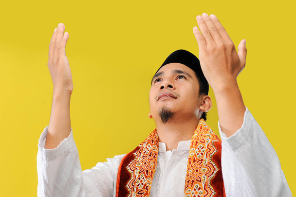 Крупным планом молодой азиат мусульманин молится, поднимая руки в мусульманском платье, низкого угла и крупным планом, изолированный на желтом фоне
 - Фото, изображение