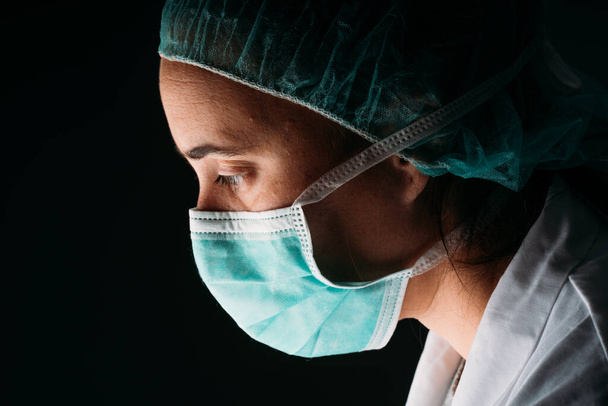 Zijdelingse jonge vrouw arts met medisch chirurgisch masker, pet en virus beschermende kleding op zwarte achtergrond met kopieerruimte. COVID-19 De coronaviruspandemie. - Foto, afbeelding