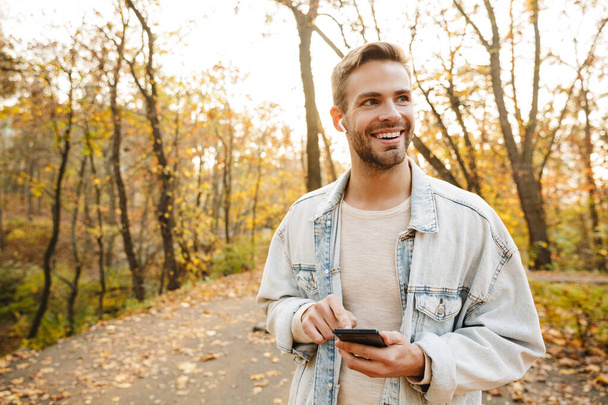 Image of handsome young caucasian man in earbuds smiling and using smartphone while walking in park - Foto, Imagem