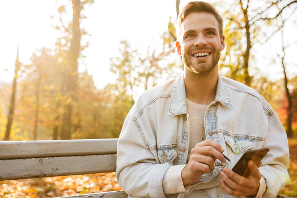 Schöner lächelnder junger Mann sitzt auf einer Bank im Herbstpark und benutzt sein Handy - Foto, Bild