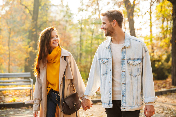 Image of attractive young caucasian couple smiling and walking through autumn park together - 写真・画像