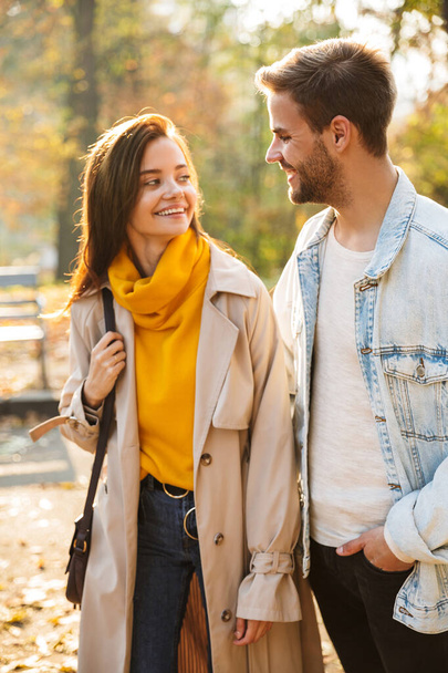 Image of attractive young caucasian couple smiling and walking through autumn park together - Fotó, kép
