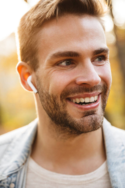 Imagen de un hombre caucásico joven guapo en chaqueta de mezclilla sonriendo y usando auriculares mientras camina en el parque
 - Foto, Imagen