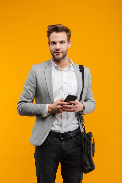 Handsome smiling young businessman wearing jacket standing isolated over yellow background, carrying bag, using mobile phone - Photo, image