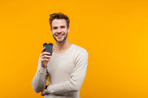Attractive happy young man wearing pullower standing isolated over yellow background, drinking takeaway coffee - 写真・画像