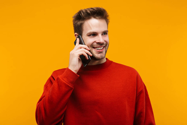 Attractive happy young man wearing pullower standing isolated over yellow background, talking on mobile phone - Fotoğraf, Görsel