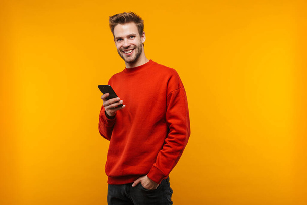 Attractive happy young man wearing pullower standing isolated over yellow background, using mobile phone - Фото, изображение