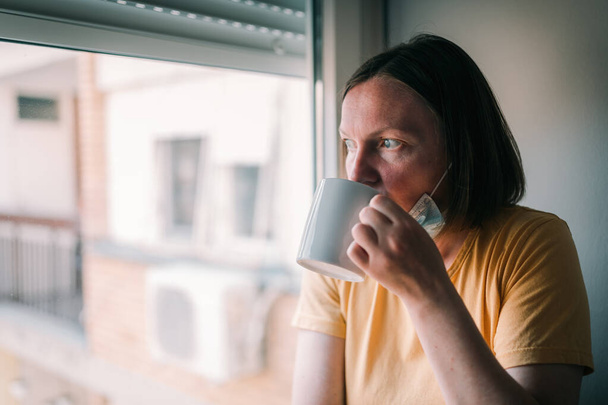 Woman in self-isolation during virus outbreak drinking coffee and looking through window. Worried female person with protective surgical mask in stay at home concept, selective focus - Photo, Image