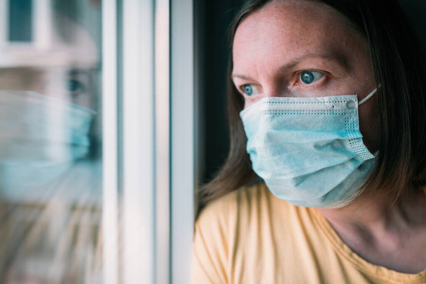 Woman in self-isolation during virus outbreak looking through window. Worried female person with protective surgical mask in stay at home concept, selective focus - Fotografie, Obrázek