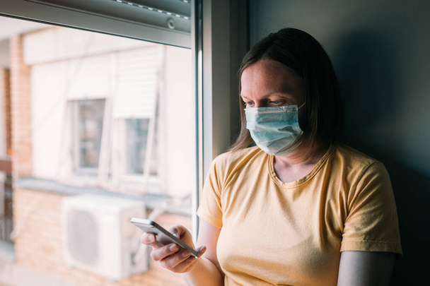 Woman in self-isolation during virus outbreak using mobile phone. Worried female person with protective surgical mask, holding smartphone in stay at home concept, selective focus - Photo, image