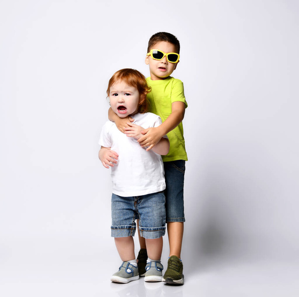 Child in sunglasses hugging his little baby brother or sister who screaming. Dressed in casual clothes, posing isolated on white - Photo, Image