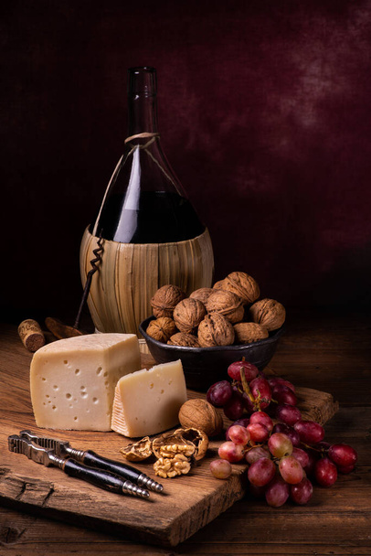 στην παλιά ξύλινη σανίδα τεμαχισμού, λίγο ώριμο κατσικίσιο τυρί, καρύδια, κόκκινα σταφύλια και στο παρασκήνιο φιάλη κρασιού - Φωτογραφία, εικόνα
