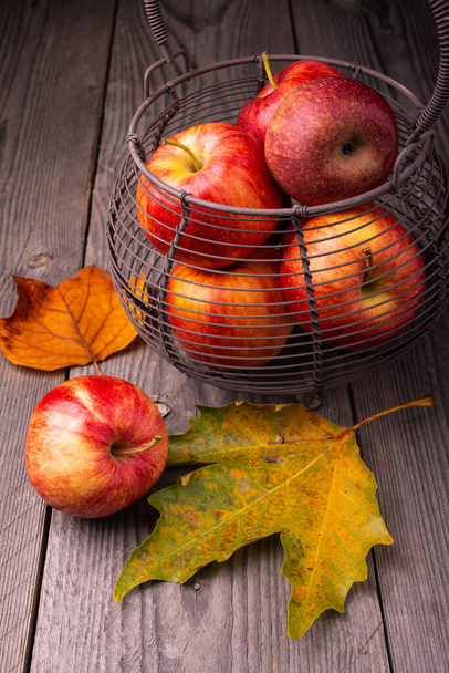 sobre la mesa de madera áspera, algunas hojas amarillentas, y una cesta de metal que contiene algunas manzanas
 - Foto, Imagen