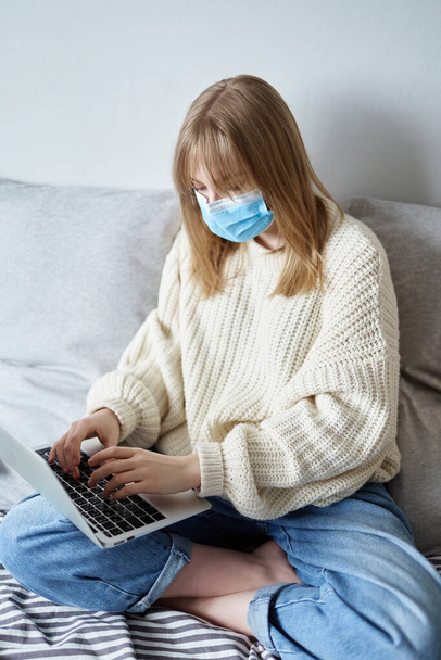 Coronavirus quarantine freelancer at remote work from home. Young teenager girl in medical mask, white sweater and jeans in isolation working at home. Concept of online studying, working, surfing or chatting during isolation period - Zdjęcie, obraz