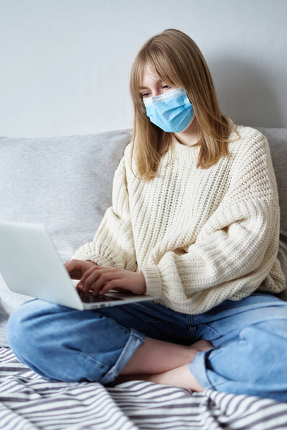 Work on remote access. Young girl in a medical mask works on a laptop at home in isolation. Concept of online studying, working, surfing or chatting during isolation period - 写真・画像