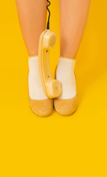 Γυναικεία πόδια στα κίτρινα παπούτσια σε κίτρινο φόντο με παλιά vintage κίτρινο ακουστικό. - Φωτογραφία, εικόνα
