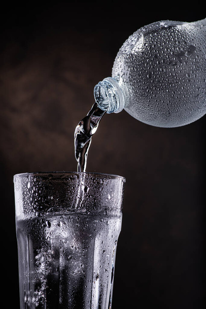 čerstvá voda nalitá z plastové láhve do skleněné kádinky na tmavém pozadí - Fotografie, Obrázek
