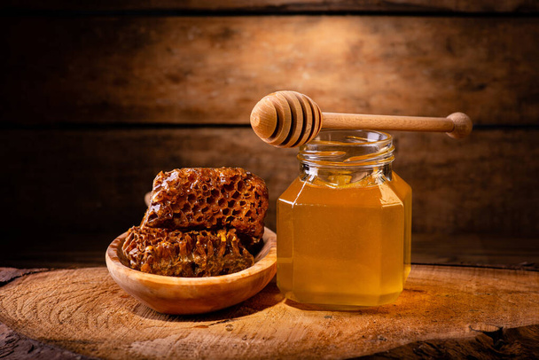 νεκρή φύση: στο τραχύ ξύλινο πάτωμα, γυάλινο βάζο με μέλι ακακίας, ξύλινη κουτάλα και κηρήθρα - Φωτογραφία, εικόνα