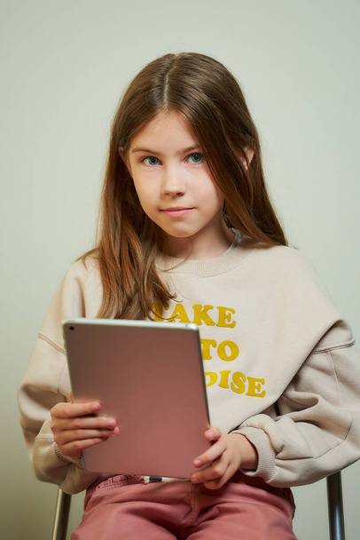 Nuori tyttö, jolla on pitkät hiukset ja joka opiskelee tabletilla. Kaunis lapsi pitelee taulutietokonetta.
. - Valokuva, kuva