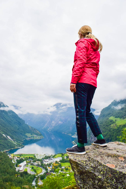 Женщина-туристка, стоящая на краю утёса возле Флайдалсьювет. Летний пейзаж небольшой деревни Гейрангер, которая находится в конце Гейрангер-фьорда. Норвегия
. - Фото, изображение