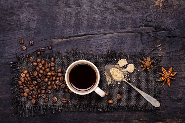 από ψηλά. Στο ωμό και σκούρο ξύλινο τραπέζι, ένα φλιτζάνι καφέ με μια κουταλιά καφέ ζάχαρη, κόκκους καφέ και γλυκάνισο αστέρι. αντίγραφο χώρου - Φωτογραφία, εικόνα