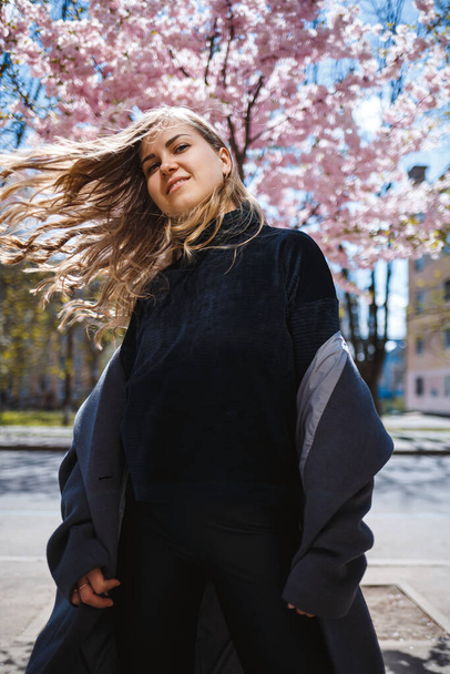 Young slender female model with long wavy hair and, dressed in a gray coat, sneakers, spinning on the street. Spring blossom trees woman girl laughs and runs to rejoice - Foto, imagen
