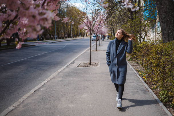 Sakura branches with flowers on a tree on the city streets. Happy woman girl in a gray palette walks along an alley with blooming sakura. Gorgeous fancy girl outdoors. Sakura tree blooming. - Valokuva, kuva