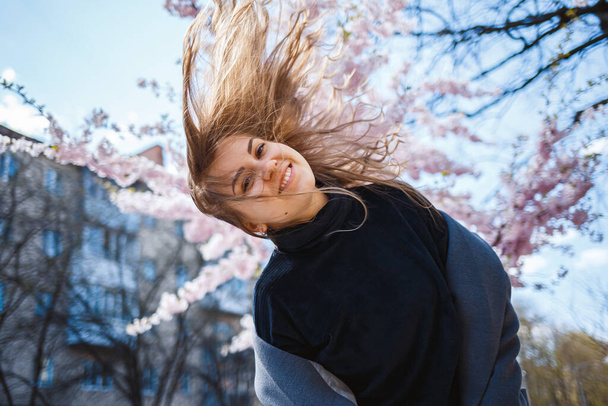 Sakura branches with flowers on a tree on the city streets. Happy woman girl spinning on the street with blooming sakura. Gorgeous fashionable girl outdoors. Sakura tree blossoms. - Photo, Image