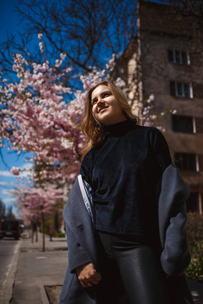 Sakura ramas con flores en un árbol en las calles de la ciudad. Mujer feliz chica en una paleta gris camina a lo largo de un callejón con sakura floreciente. Hermosa chica elegante al aire libre. Sakura árbol floreciendo
. - Foto, Imagen