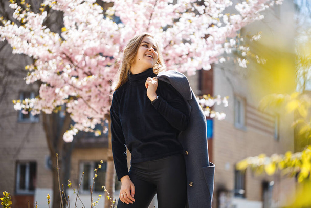 Os ramos de Sakura com flores em uma árvore nas ruas de cidade. Menina mulher feliz em uma paleta cinza caminha ao longo de um beco com sakura florescendo. Menina fantasia lindo ao ar livre. Árvore Sakura florescendo
. - Foto, Imagem