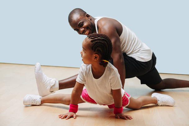 Pai negro feliz e sua filhinha se exercitando em quarentena juntos, esticando as pernas em meia-divisão
. - Foto, Imagem
