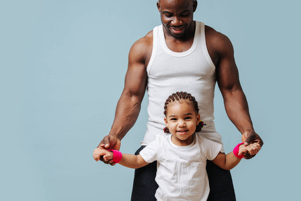 Happy fit family in isolation. Black father and daughter in sportive clothes showing up muscles over blue background. - Fotoğraf, Görsel