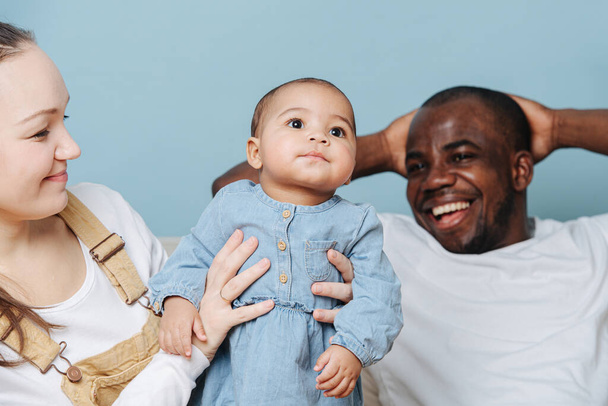 Happy mixed race family posing for a family photo in a studio. Black father, white moher and their jolly infant daughter over blue background. - Zdjęcie, obraz