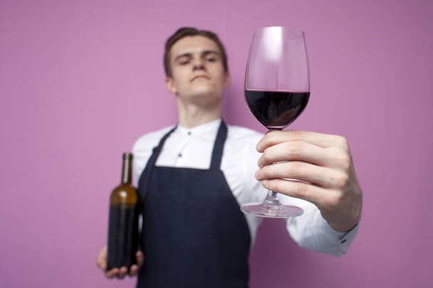 giovane cameriere professionista in uniforme regala un bicchiere di vino rosso su sfondo rosa, il sommelier propone di bere vino, primo piano
 - Foto, immagini