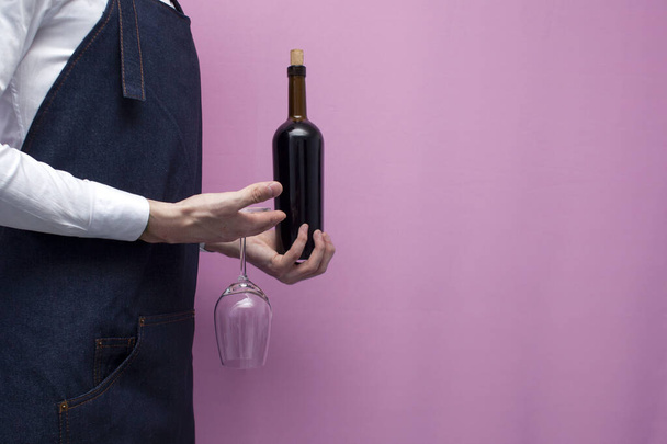 νεαρός επαγγελματίας σερβιτόρος με στολή κρατά ένα μπουκάλι κόκκινο κρασί και ένα ποτήρι σε ροζ φόντο, κοντινό πλάνο - Φωτογραφία, εικόνα