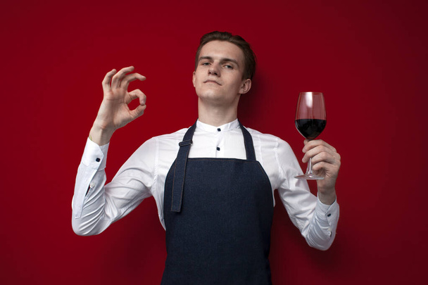 νεαρός επαγγελματίας οινοχόος με ένα ποτήρι κόκκινο κρασί δείχνει εντάξει σε ένα κόκκινο φόντο, ένας εμπειρογνώμονας τύπος δίνει μια καλή εκτίμηση του κρασιού - Φωτογραφία, εικόνα