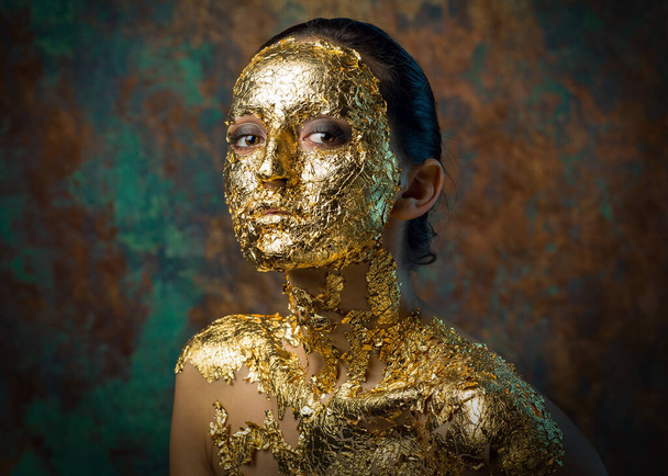 Girl with a mask on her face made of gold leaf. Gloomy studio portrait of a brunette on an abstract background. - 写真・画像