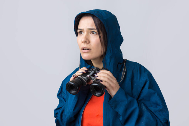 A tourist girl in a blue raincoat holds binoculars in her hands and looks into the distance, spies. - Foto, Imagem