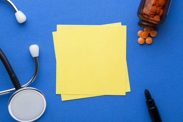 Nota gialla vuota con stetoscopio, pillole e vari articoli di cancelleria su sfondo blu con spazio per il testo. Concetto medico
 - Foto, immagini