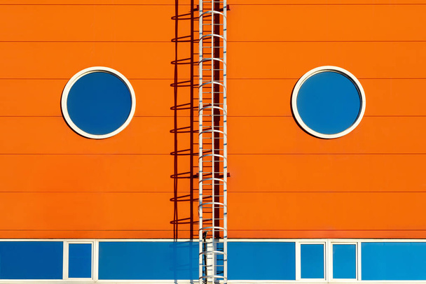 Fragment nowoczesnej pomarańczowej fasady budynku przemysłowego, magazynu lub centrum wystawowego z dwoma symetrycznymi okrągłymi oknami i metalowymi schodami. Stowarzyszenie Graficzne z twarzą osoby - Zdjęcie, obraz