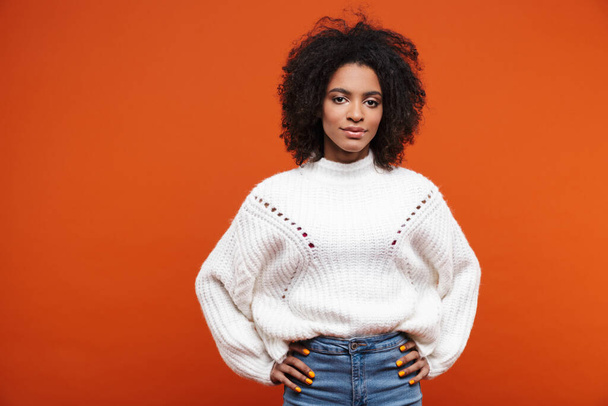 Beautiful smiling young african woman wearing sweater standing islolated over red background, hands on hips - Фото, зображення