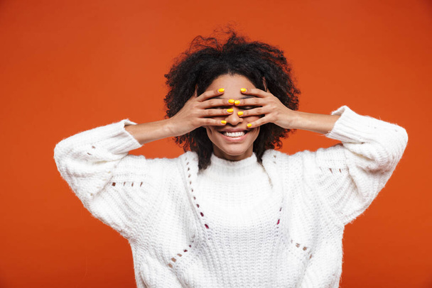 Beautiful cheerful young african woman wearing sweater standing islolated over red background, covering eyes - Foto, afbeelding