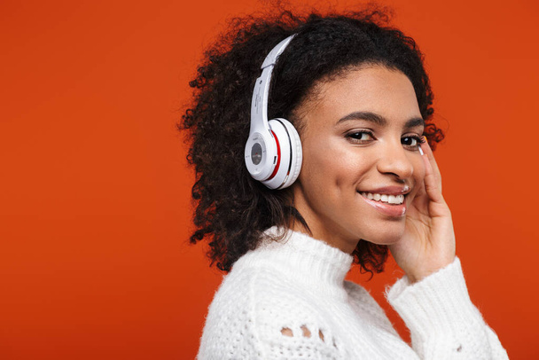 赤の背景の上に隔離されたワイヤレスヘッドフォンで音楽を聴く陽気な若いアフリカの女性 - 写真・画像