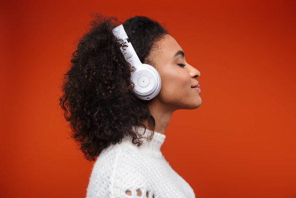 Side view of a cheerful young african woman listening to music with wireless headphones isolated over red background - Foto, Imagen