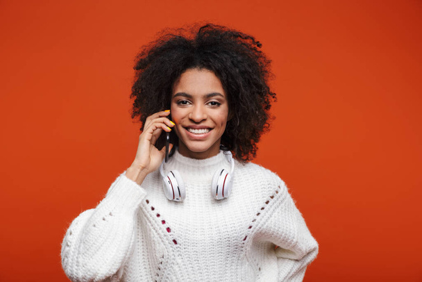 Attractive young african woman wearing sweater talking on mobile phone isolated over red background - Фото, зображення