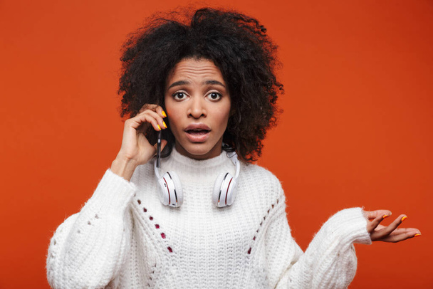 Attractive confused young african woman wearing sweater talking on mobile phone isolated over red background - Fotó, kép