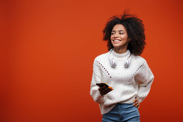 魅力的な笑顔若いアフリカ人女性を身に着けていますセーターを保持携帯電話赤の背景に隔離 - 写真・画像