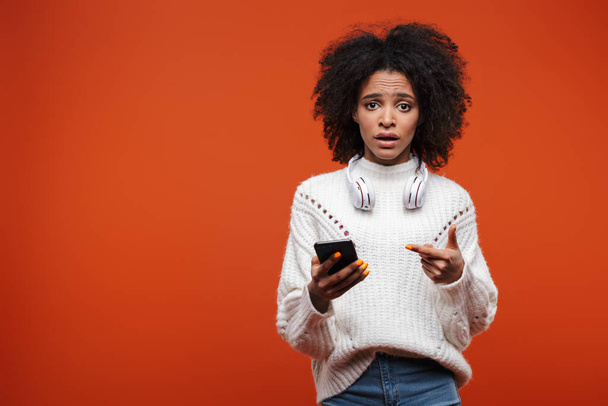 Attractive shocked young african woman wearing sweater holding mobile phone isolated over red background - Photo, Image