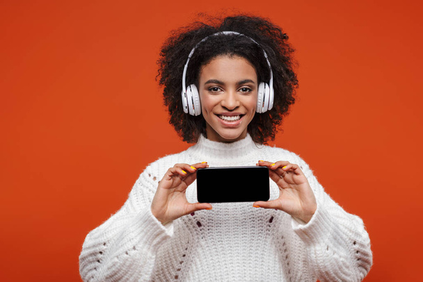 Cheerful attractive young african woman wearing wireless headphones standing isolated over red background, showing blank screen mobile phone - Foto, imagen